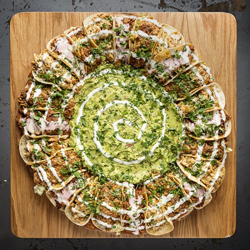 A circular platter of tacos arranged in a spiral, topped with lettuce, sauce, and garnished with cilantro.