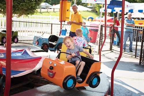A young boy joyfully rides a colorful amusement park car, with other children and adults in the background.