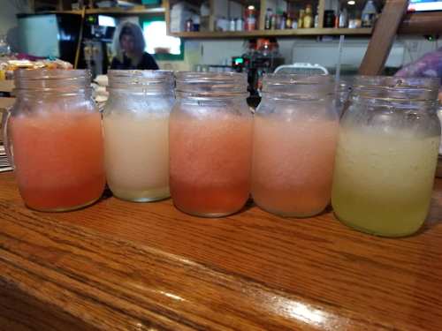 Five mason jars filled with colorful slushy drinks, arranged on a wooden bar counter.