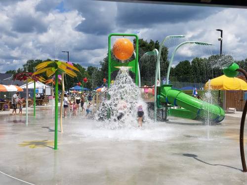 A vibrant water park scene with kids playing in water, colorful slides, and splash features under a cloudy sky.