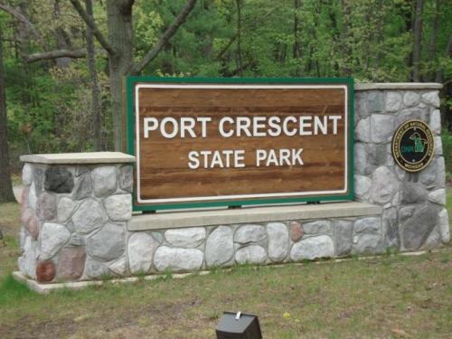 Sign for Port Crescent State Park, featuring wooden and stone design, surrounded by trees and greenery.