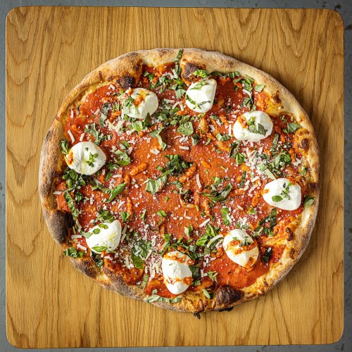 A round pizza topped with marinara sauce, fresh basil, mozzarella balls, and grated cheese on a wooden surface.