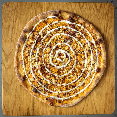 A circular pizza topped with cheese, diced toppings, and a spiral of white sauce on a wooden surface.
