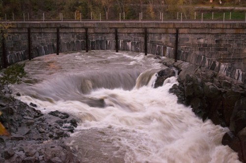 A rushing river flows under a stone bridge, surrounded by rocky banks and trees in the background.