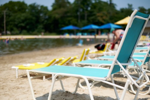 Lounge chairs in turquoise and yellow on a sandy beach near a calm water body, with umbrellas in the background.