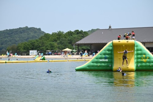 A water park scene with people on an inflatable slide and others swimming in a lake, surrounded by trees and a building.