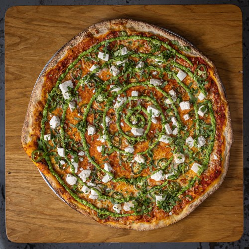 A circular pizza topped with spiral patterns of green peppers, herbs, and chunks of white cheese on a wooden surface.