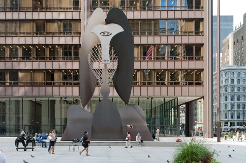 A large abstract sculpture stands in front of a modern building, with people walking and sitting nearby.