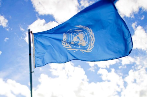A blue flag with the United Nations emblem waving against a cloudy sky.