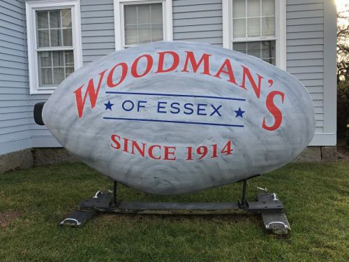 A large, oval sign reading "Woodman's of Essex Since 1914" in red and blue, displayed on a grassy area.