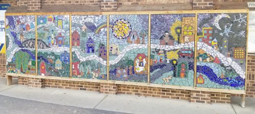 Colorful mosaic mural depicting a whimsical landscape with houses, trees, and a winding river.