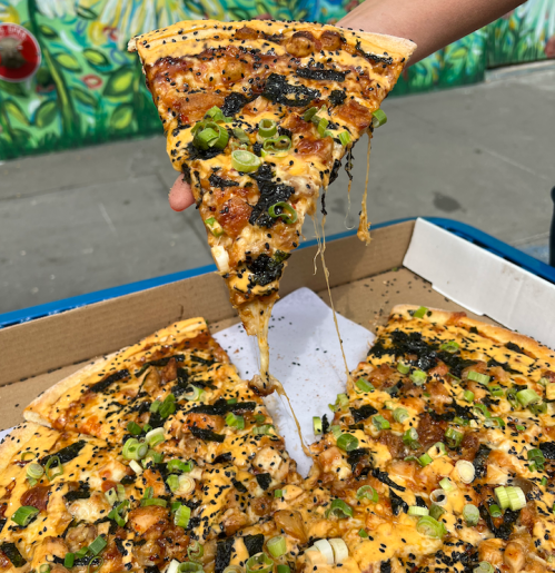 A hand holds a slice of cheesy pizza topped with green onions, with a box of pizza in the background.