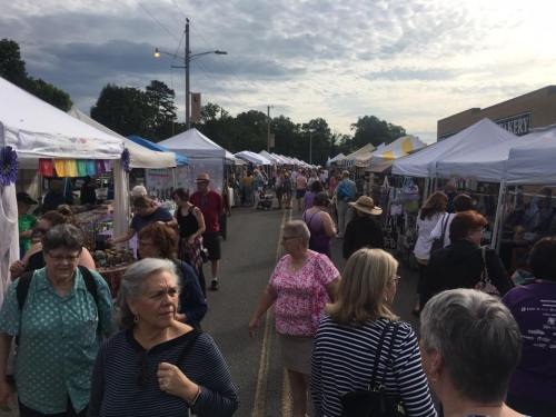 A bustling outdoor market with numerous tents, shoppers browsing, and a cloudy sky overhead.