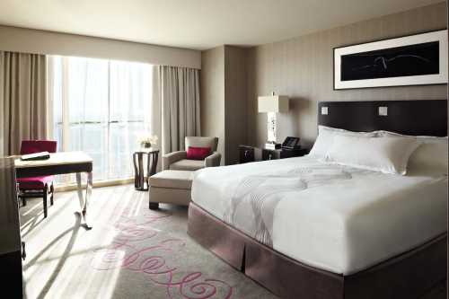 A modern hotel room featuring a large bed, a chair, a desk, and large windows with natural light.