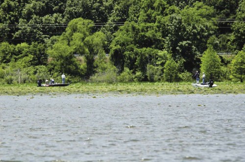 Two boats on a lake near a lush green shoreline, with people fishing among the lily pads.
