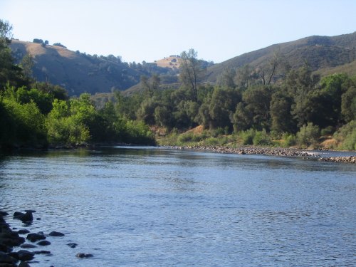 A serene river flows through a lush landscape, surrounded by hills and trees under a clear blue sky.