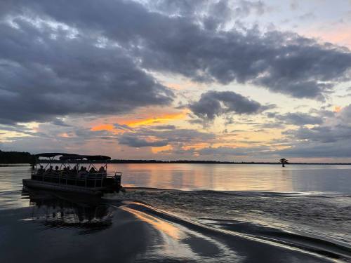 A boat with passengers glides across a calm lake at sunset, with colorful clouds reflecting on the water.