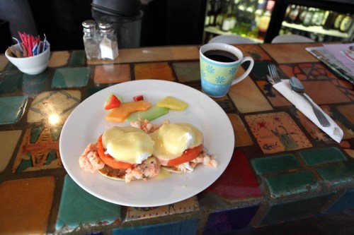 A plate of eggs Benedict with shrimp and tomato, accompanied by fruit and a cup of coffee on a colorful table.