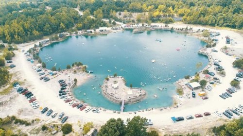 Aerial view of a heart-shaped lake surrounded by trees, with cars parked nearby and people enjoying the water.