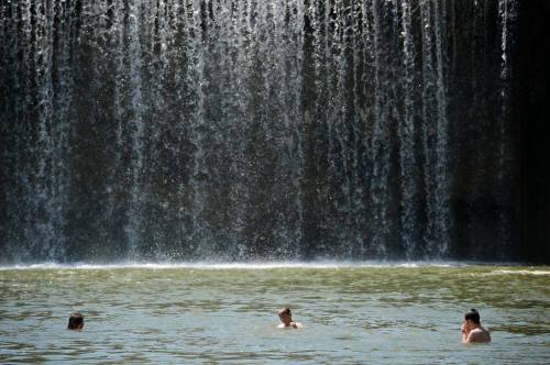 Three people wade in a pool beneath a cascading waterfall, surrounded by natural scenery.