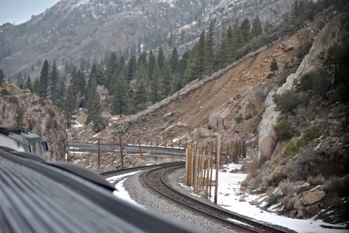 A train track curves through a snowy landscape with mountains and pine trees in the background.