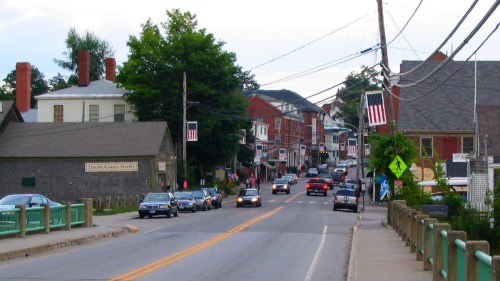 A small town street lined with buildings, cars, and American flags, with power lines overhead and trees in the background.