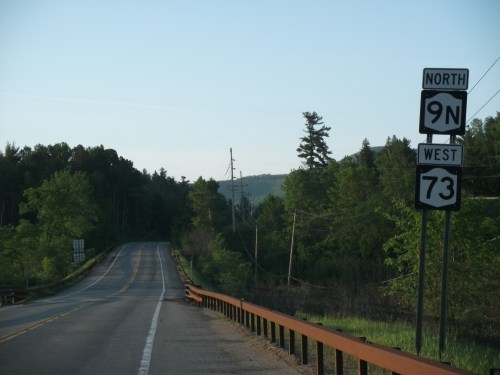 A rural road sign indicating North 9N and West 73, surrounded by greenery and distant hills.