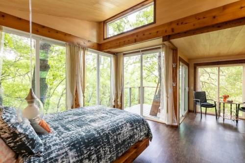 Cozy bedroom with large windows overlooking greenery, featuring a bed, wooden accents, and a small dining area.