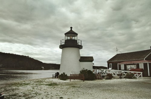 A white lighthouse beside a calm waterway, with a cloudy sky and a nearby building and benches.