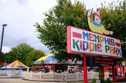 Colorful sign for Memphis Kiddie Park, featuring a clown, with amusement rides and trees in the background.