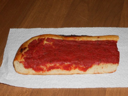 A rectangular piece of bread topped with a thick layer of red tomato sauce, resting on a paper towel.