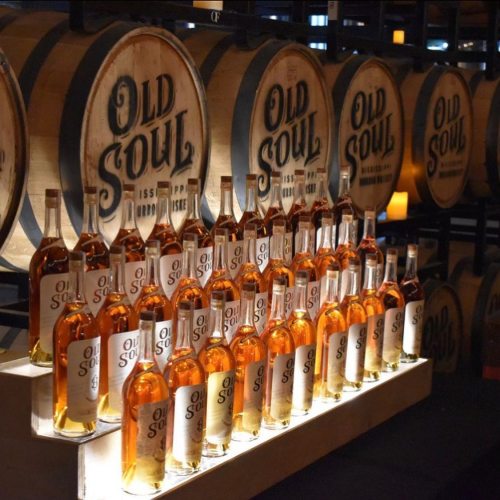 A display of bottles labeled "Old Soul" in front of wooden barrels, showcasing a warm amber liquid.