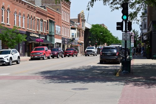A street scene featuring parked cars, shops, and green traffic lights on a sunny day.