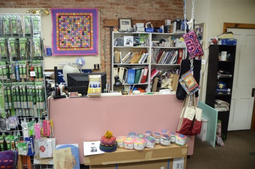 A craft store interior featuring a pink counter, shelves of supplies, and colorful fabric displays.