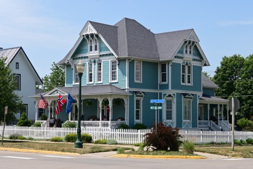 A large, blue Victorian-style house with white trim, surrounded by greenery and flags, on a sunny day.