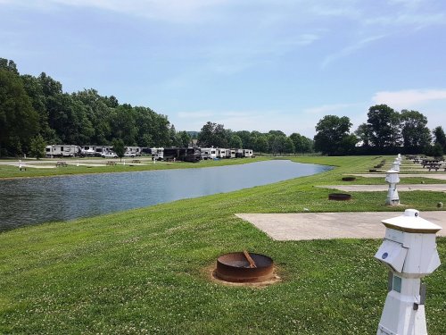 A serene RV park by a calm waterway, surrounded by green trees and grassy areas, with picnic spots and fire pits.