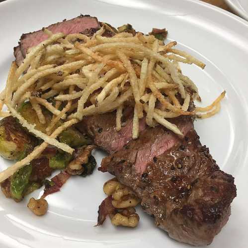 A plated steak topped with crispy fried onions, served with Brussels sprouts and nuts on a white dish.