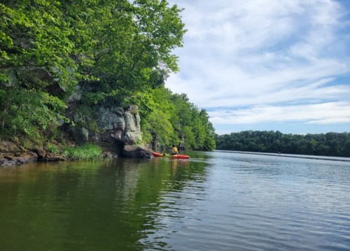 Two kayakers near a rocky shore on a calm lake, surrounded by lush green trees under a blue sky.