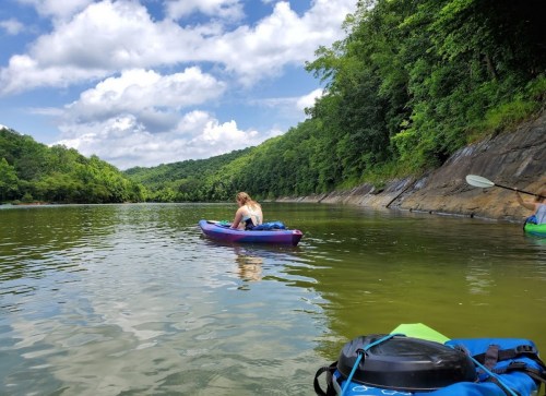 A person in a kayak paddles on a calm river surrounded by lush green hills and a partly cloudy sky.
