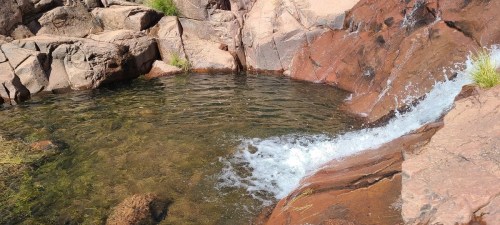 A serene natural pool with clear water, surrounded by rocky terrain and a small waterfall cascading over the stones.