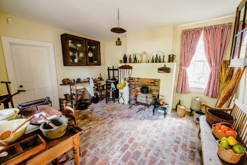 A cozy vintage kitchen with a brick fireplace, wooden furniture, and various kitchenware displayed on shelves.