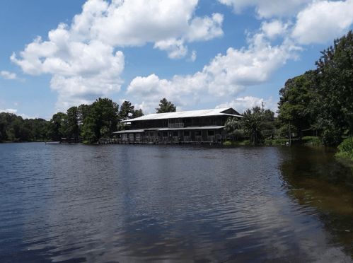 A serene lake scene with a wooden building, surrounded by trees and fluffy clouds in a blue sky.
