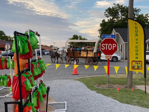 A horse-drawn wagon passes a stop sign near a colorful display of bags and a yellow banner for a local event.