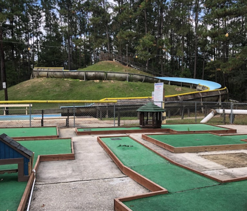 A mini-golf course with green turf and wooden borders, featuring a large slide and a hill in the background.
