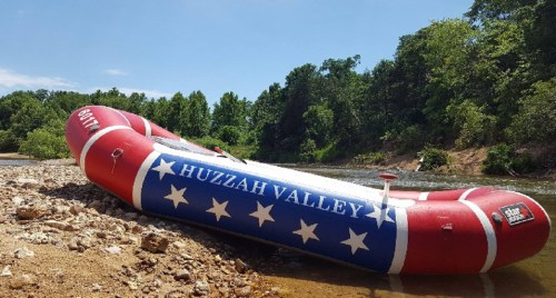 A red, white, and blue inflatable raft labeled "Huzzah Valley" rests on a rocky riverbank under a clear blue sky.