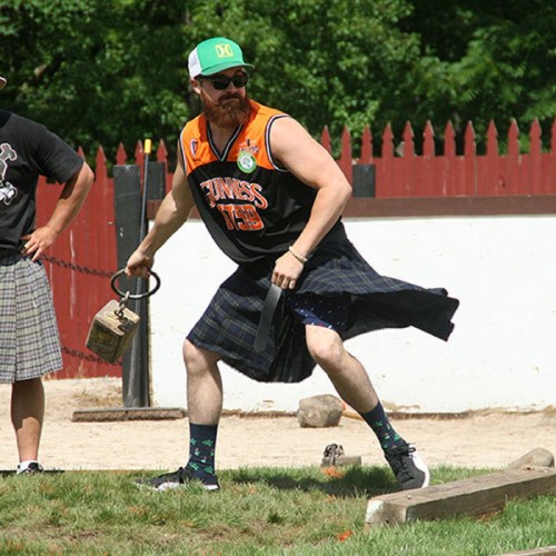 A man in a kilt and tank top throws a heavy weight, wearing a cap and colorful socks, with a grassy background.