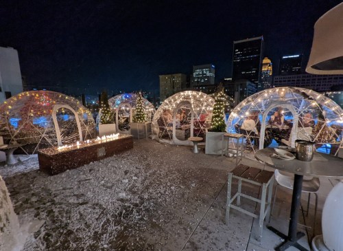 A rooftop scene with illuminated igloo-like structures, surrounded by snow and city skyline at night.