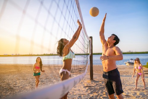 A beach volleyball game at sunset, with two players jumping to hit the ball while two others watch.