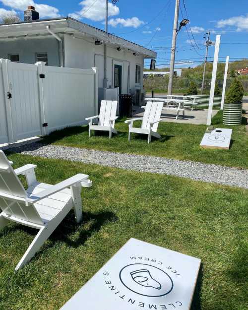 A sunny outdoor area with white adirondack chairs and cornhole boards near a creamery building. Green grass and blue sky.