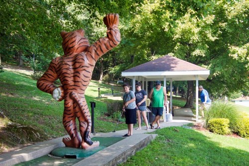 A large tiger statue waves as people play mini-golf nearby, surrounded by trees and a gazebo in the background.